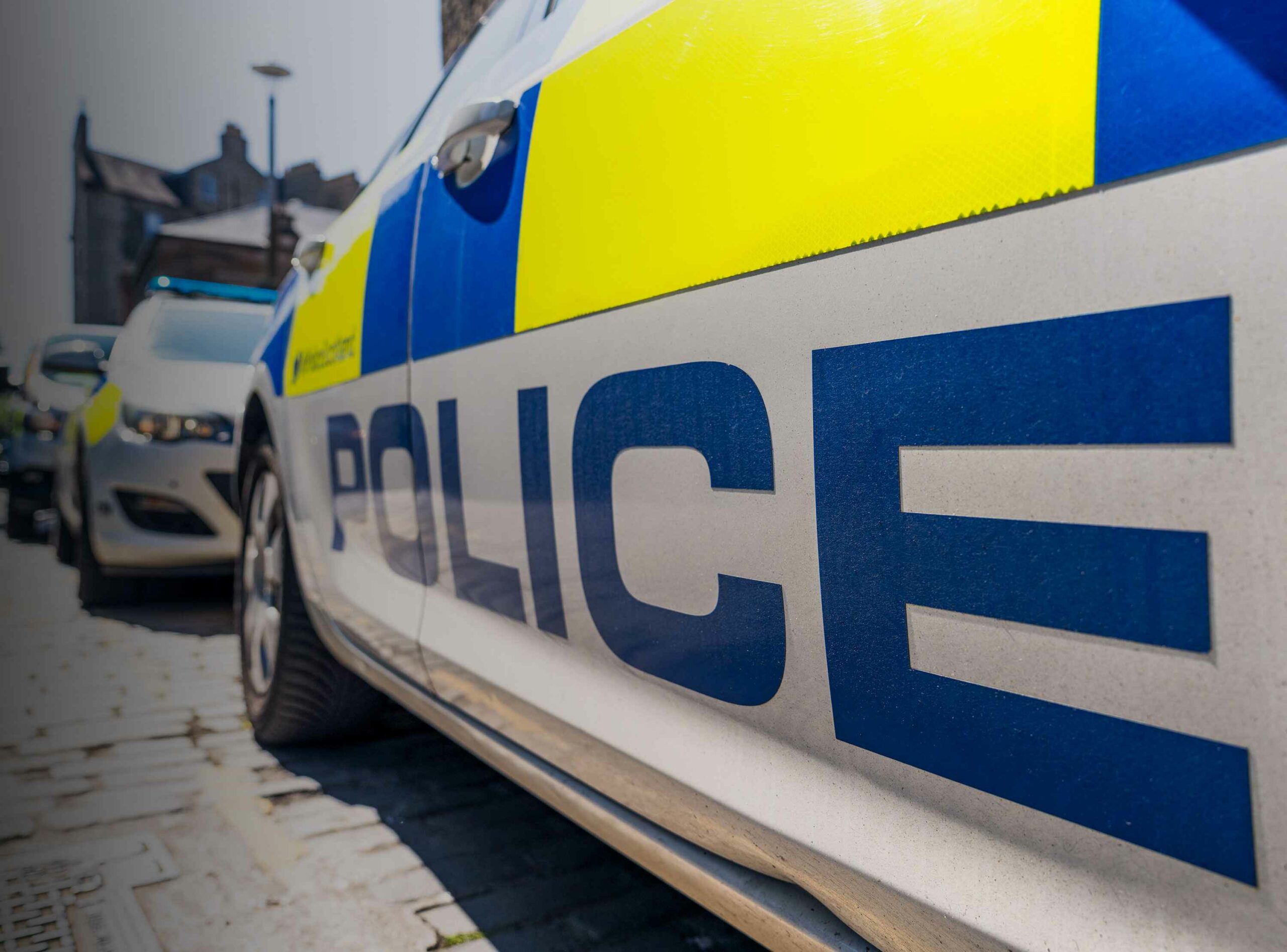 The word 'Police' below the fluorescent decals on a police car on a UK street, with other police vehicles in the distance.