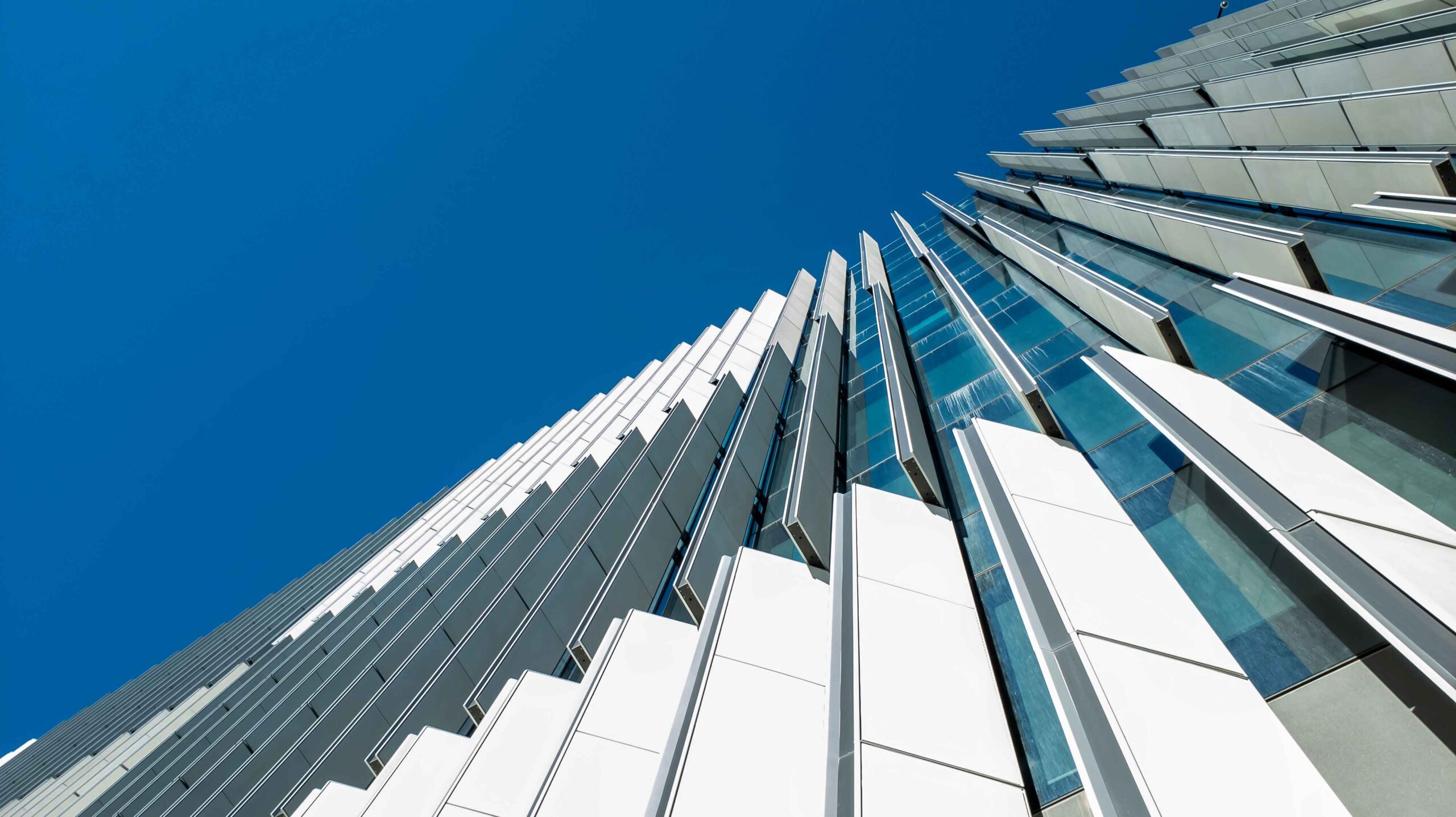 Upward view from street level of a tall modern office building exterior. Against a blue sky. Adjustable external slats to control incoming natural sunlight for internal light and temperature control.
