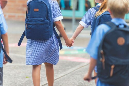 School children walking away. They are wearing uniforms and carrying backpacks. They are having a race. Multi ethnic group with Caucasian and Aboriginal children. Rear view. girls are holding hands
